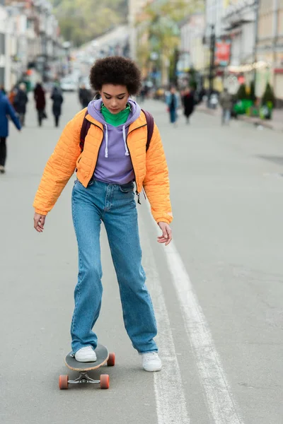 Junge Afroamerikanerin mit Rucksack reitet Longboard auf urbaner Straße — Stockfoto