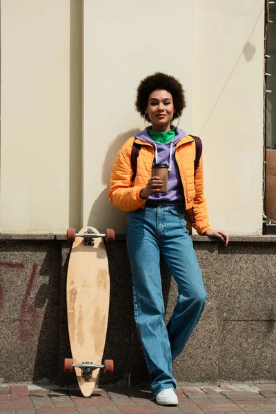 Smiling african american woman with paper cup looking at camera near building and longboard — Stock Photo