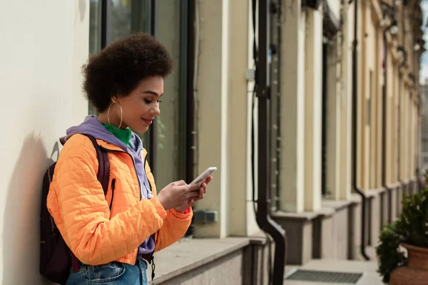 Femme afro-américaine souriante avec sac à dos en utilisant un smartphone près du bâtiment — Photo de stock