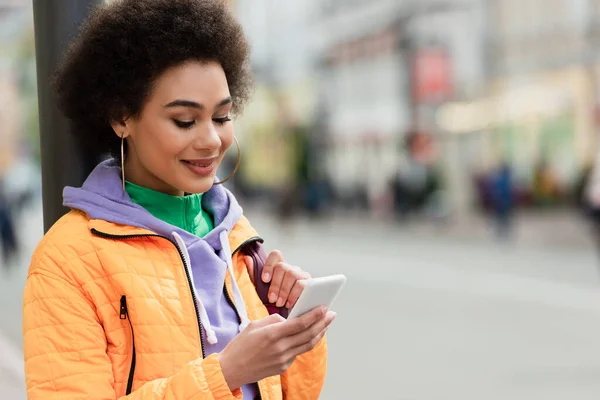 Femme afro-américaine avec sac à dos utilisant un téléphone portable et souriant à l'extérieur — Photo de stock