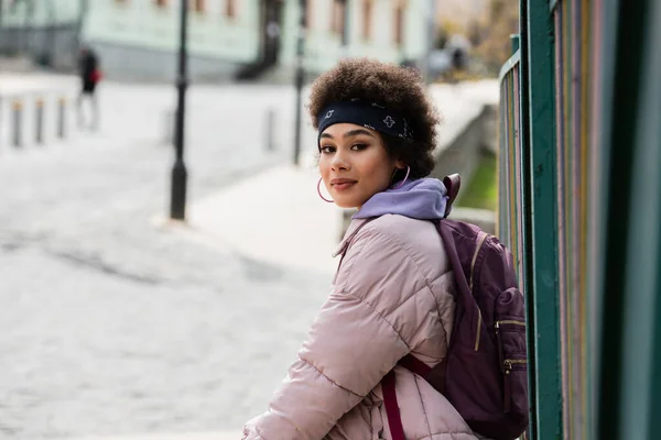 Jovem afro-americana com mochila olhando para a câmera perto de cerca na rua urbana — Fotografia de Stock