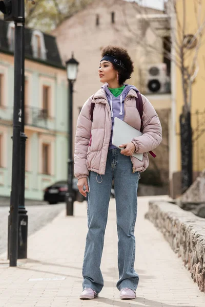 Femme afro-américaine élégante tenant un ordinateur portable tout en marchant dans la rue urbaine — Photo de stock