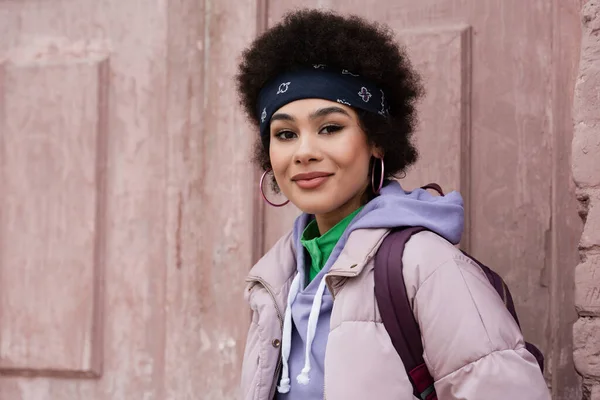 Femme afro-américaine en veste et bandana regardant la caméra près de la porte du bâtiment — Photo de stock