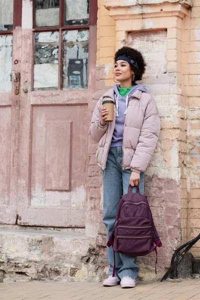 Stilvolle afrikanisch-amerikanische Frau mit Coffee to go und Rucksack in der Nähe von Altbau — Stockfoto