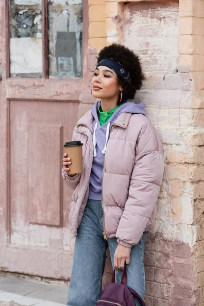 Pretty african american woman with backpack holding coffee to go near old building — Stock Photo