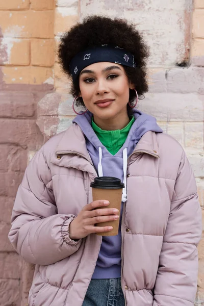 Femme afro-américaine élégante tenant une tasse en papier et regardant la caméra à l'extérieur — Photo de stock