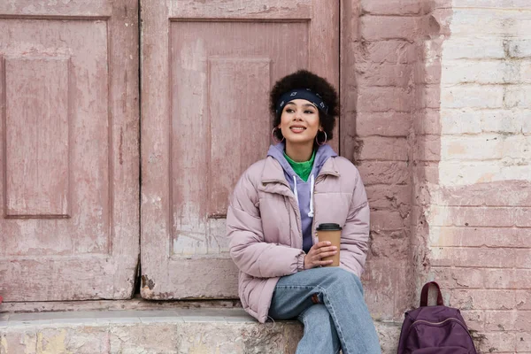 Feliz mulher afro-americana segurando café para ir perto de mochila e prédio antigo — Fotografia de Stock