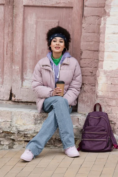 Sonriente mujer afroamericana sosteniendo café para ir cerca de la mochila en la calle urbana - foto de stock