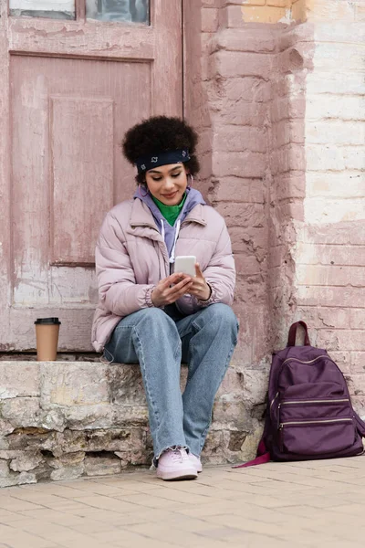 Усміхнена афро-американська жінка, користуючись смартфоном біля кави, йде і рюкзак на міській вулиці. — стокове фото