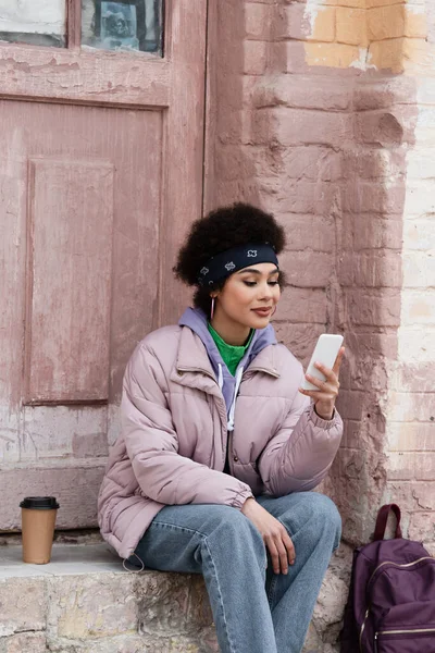 Stylish african american woman using smartphone near paper cup and backpack on stair of building — Stock Photo