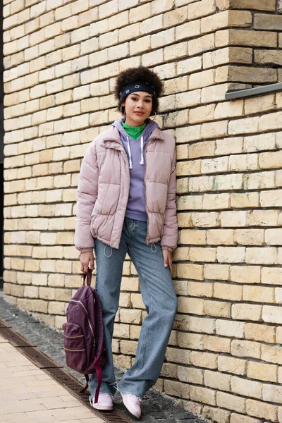 Africano mulher americana com mochila olhando para a câmera perto da parede de tijolo — Fotografia de Stock