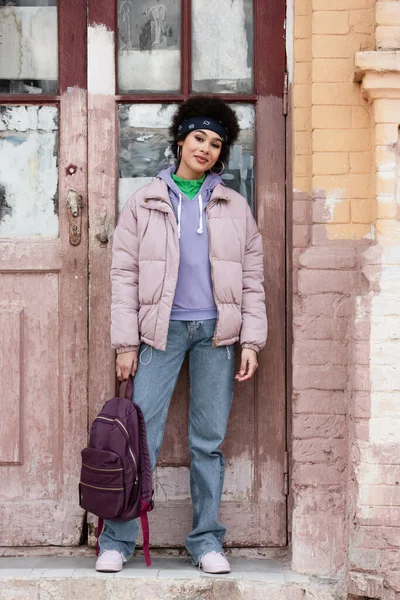 Afroamerikanerin mit Rucksack steht in der Nähe von Altbau mit Holztür — Stockfoto