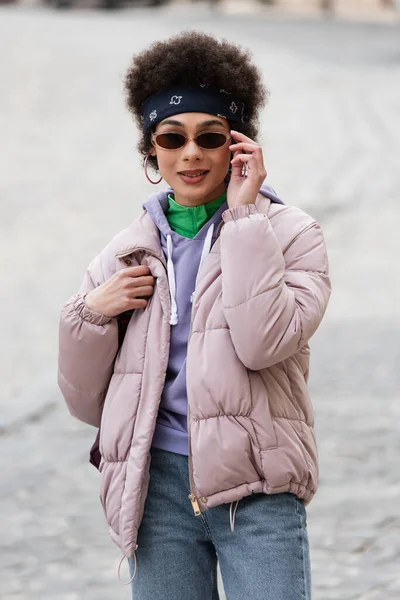 Mujer afroamericana en gafas de sol sosteniendo mochila y sonriendo en la calle urbana - foto de stock