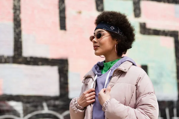 Femme afro-américaine en lunettes de soleil ajustant la veste à l'extérieur — Photo de stock
