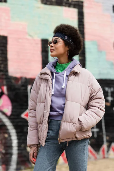 African american woman in sunglasses holding hand in pocket of jacket on urban street — Stock Photo