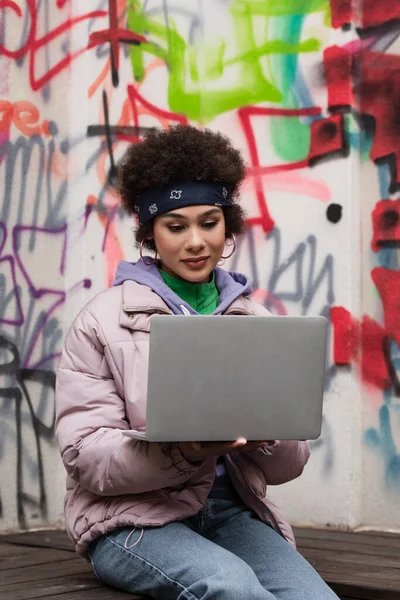 Молодая американка из Африки пользуется ноутбуком рядом с граффити на открытом воздухе — стоковое фото