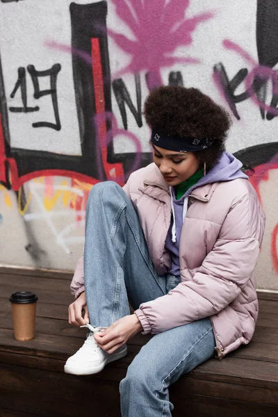 Mujer afroamericana atando cordones de zapatos cerca de la taza de papel en el banco y graffiti en la pared - foto de stock