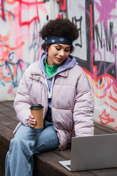 Élégant pigiste afro-américain avec tasse en papier à l'aide d'un ordinateur portable sur un banc près de graffitis à l'extérieur — Photo de stock