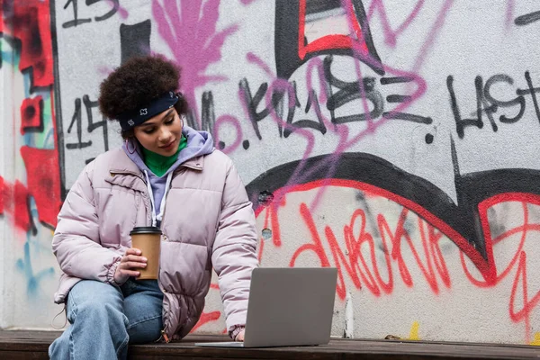 Femme afro-américaine tenant du café pour aller et en utilisant un ordinateur portable près de graffitis à l'extérieur — Photo de stock