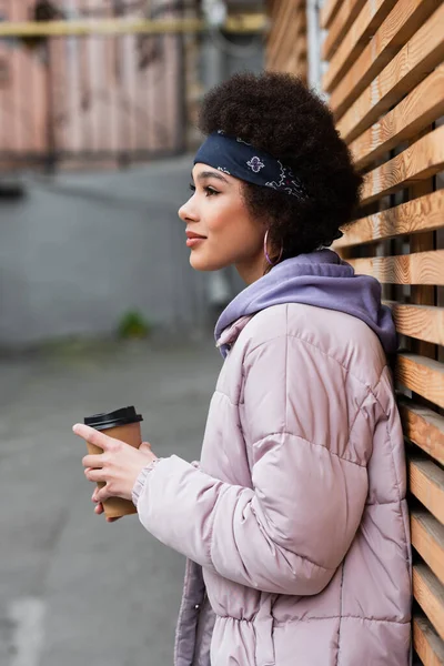 Vista lateral da mulher americana africana em bandana e jaqueta segurando copo de papel perto de cerca de madeira — Fotografia de Stock