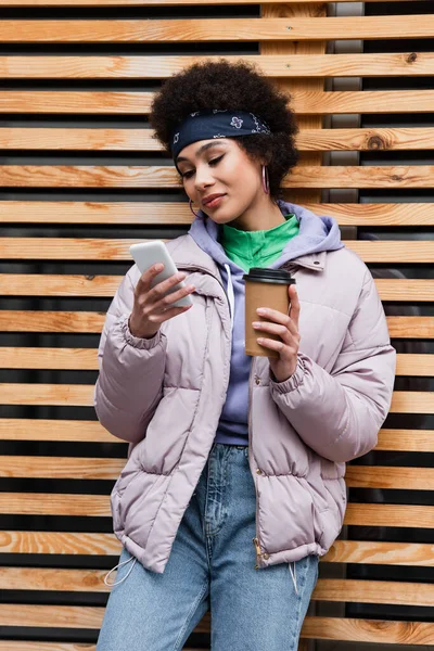 Donna afroamericana con caffè per andare con il cellulare vicino alla recinzione di legno — Foto stock