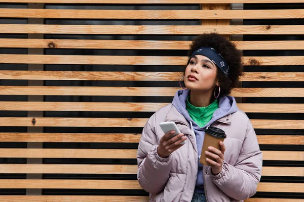 Femme afro-américaine avec café et smartphone debout près d'une clôture en bois à l'extérieur — Photo de stock