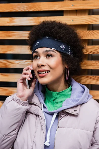 Mujer afroamericana sonriente hablando en un teléfono inteligente cerca de una cerca de madera borrosa - foto de stock