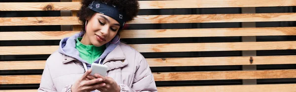 Femme afro-américaine utilisant un smartphone près d'une clôture en bois à l'extérieur, bannière — Photo de stock