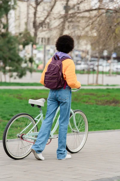 Vue arrière de la femme afro-américaine avec sac à dos tenant le vélo et marchant à l'extérieur — Photo de stock