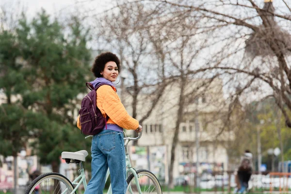 Africano americano mulher em jaqueta de pé perto de bicicleta ao ar livre — Fotografia de Stock