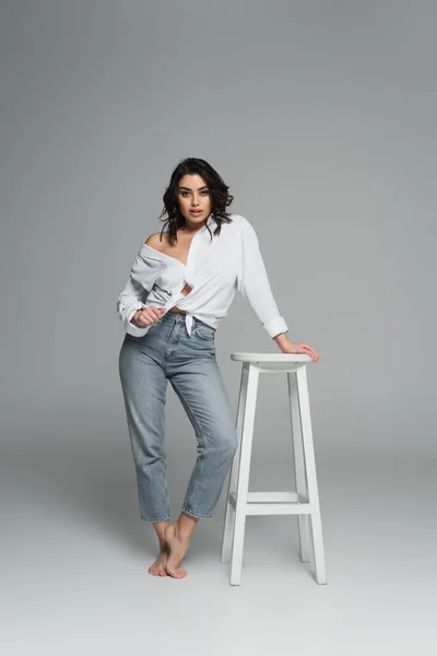 Чуттєва жінка в сорочці і джинсах, що стоїть біля стільця на сірому фоні — стокове фото