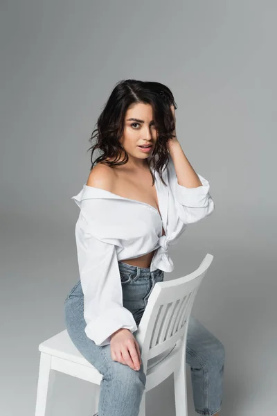 Sensual mujer en camisa ajustando el pelo en silla sobre fondo gris - foto de stock