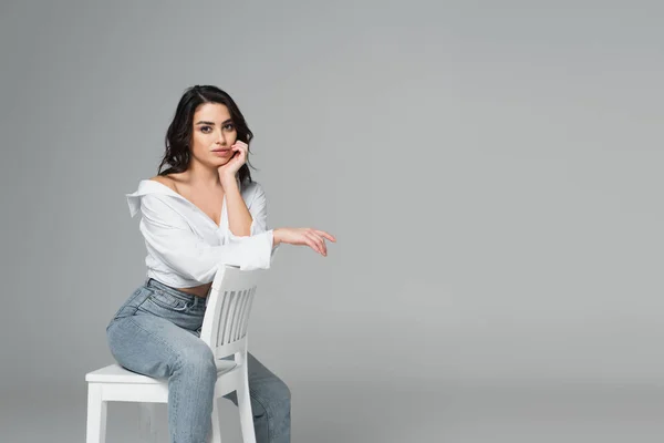 Hübsche Frau blickt in die Kamera, während sie auf weißem Stuhl isoliert auf grau sitzt — Stockfoto