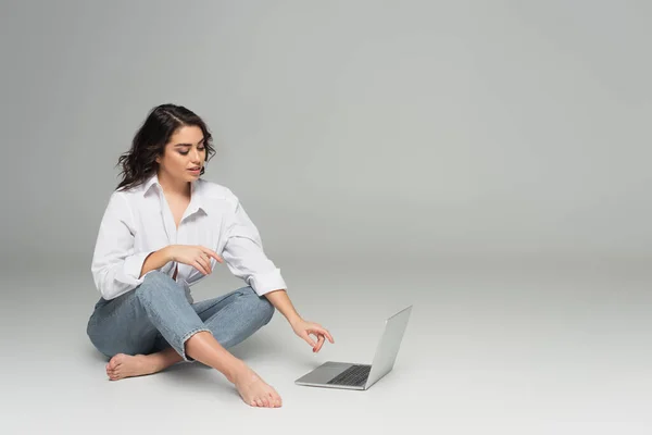 Femme souriante en chemise et jeans utilisant un ordinateur portable sur fond gris — Photo de stock