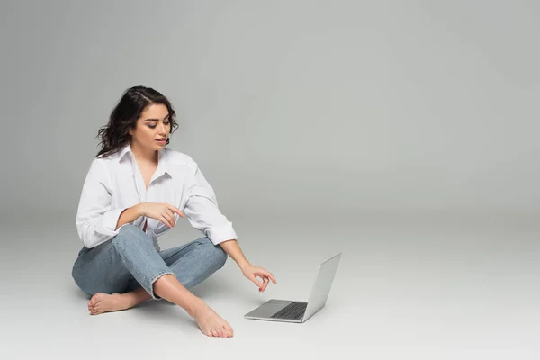 Donna sorridente in camicia e jeans con computer portatile su sfondo grigio — Foto stock