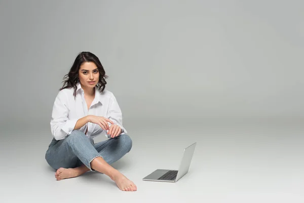 Mujer sonriente en jeans y camisa mirando a la cámara cerca de la computadora portátil sobre fondo gris - foto de stock