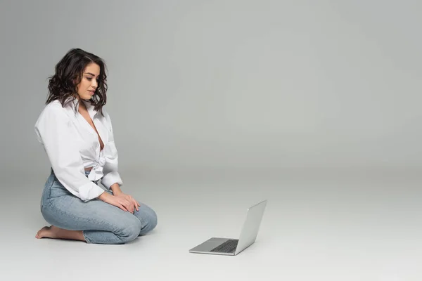 Sexy mujer morena mirando a la computadora portátil mientras está sentado sobre fondo gris - foto de stock