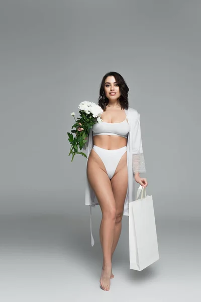 Smiling woman in lingerie holding flowers and shopping bag on grey background — Stock Photo