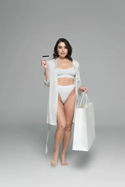 Mulher sensual em robe de cetim e lingerie segurando cartão de crédito e saco de compras em fundo cinza — Fotografia de Stock