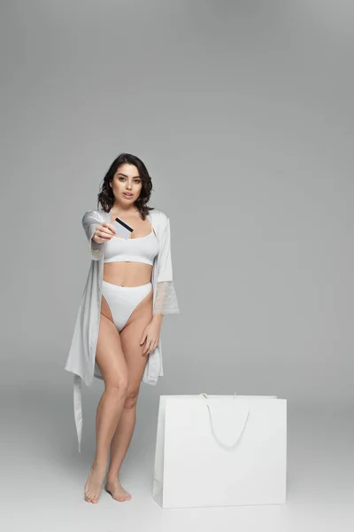 Modello seducente in lingerie che mostra carta di credito vicino shopping bag su sfondo grigio — Foto stock