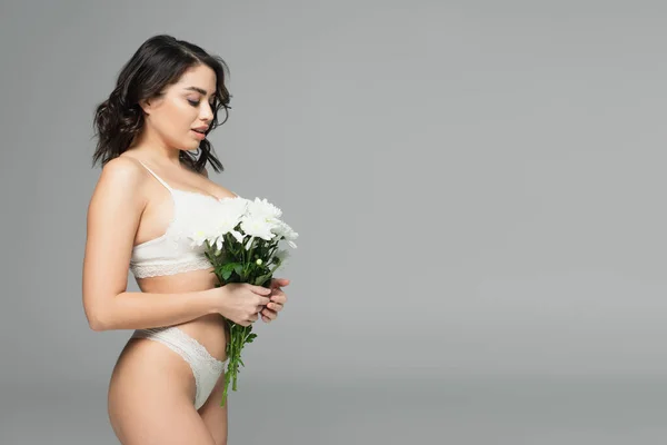 Vista lateral de mujer sensual en ropa interior sosteniendo crisantemos blancos aislados en gris - foto de stock