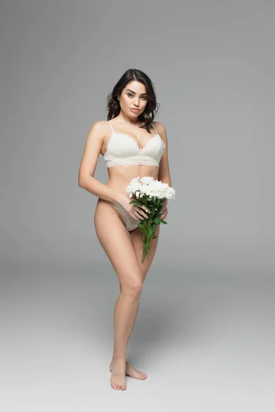 Mulher bonita morena em renda lingerie segurando flores e olhando para a câmera no fundo cinza — Fotografia de Stock