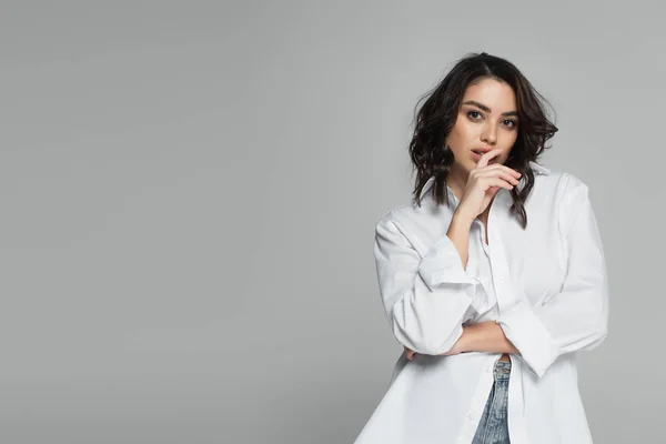 Mujer morena en camisa blanca tocando labios aislados en gris - foto de stock