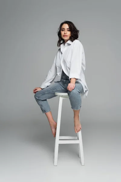 Pretty woman in jeans and shirt on chair on grey background — Stock Photo