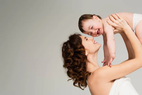 Alegre madre con hombros desnudos sosteniendo en brazos bebé niño aislado en gris - foto de stock