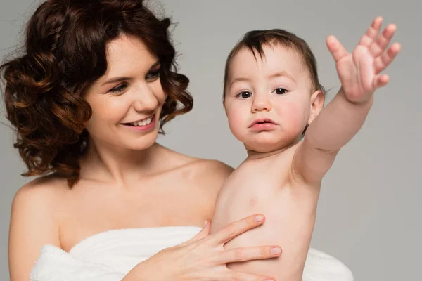 Lächelnde lockige Mutter mit nackten Schultern, die ihren Sohn mit ausgestreckter Hand auf grau isoliert in den Armen hält — Stockfoto