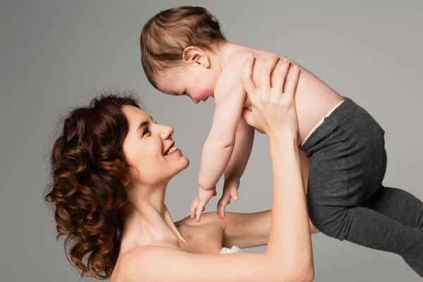 Glückliche Mutter mit nackten Schultern, die Sohn in Babystrumpfhosen auf grau isoliert in den Armen hält — Stockfoto