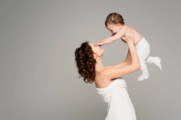 Junge in Strumpfhosen berührt Gesicht der glücklichen Mutter in Handtuch isoliert auf grau — Stockfoto