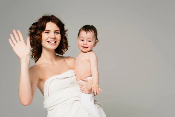 Feliz madre en toalla sosteniendo en brazos hijo en medias de bebé y agitando la mano aislada en gris - foto de stock