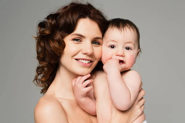 Curly mother with bare shoulders smiling and holding in arms baby boy isolated on grey — Stock Photo